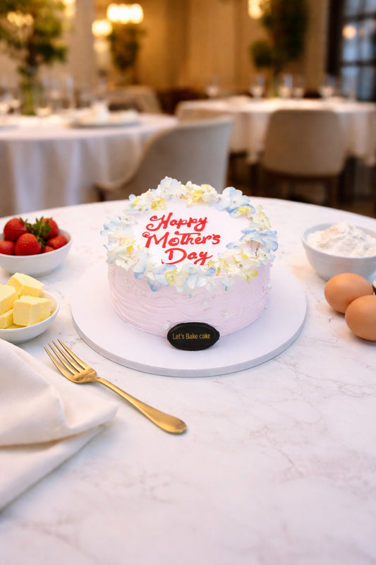 A luxury soft-pink textured cake with 'Happy Mother's Day' written in red, decorated with a blue and white floral crown, set on a marble table with strawberries and gold cutlery in Abu Dhabi, by Let's Bake Cake.