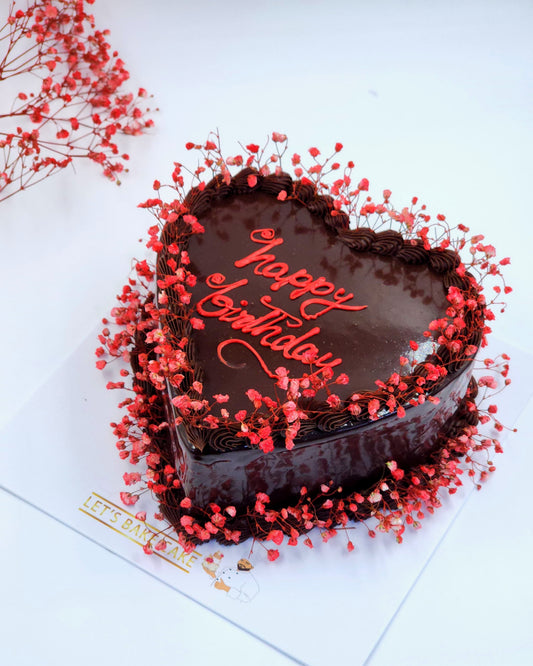 A heart-shaped dark chocolate glossy cake decorated with red dried flowers and 'Happy Birthday' calligraphy by Let's Bake Cake Abu Dhabi