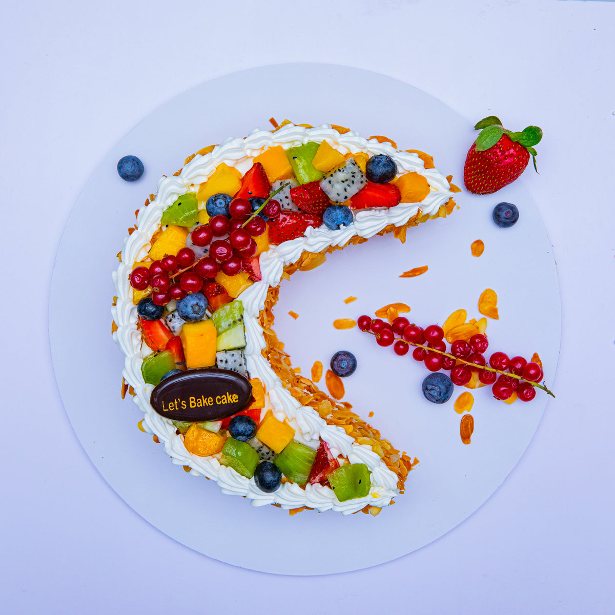 A luxury crescent moon-shaped cake topped with fresh dragon fruit, kiwi, mango, and berries, decorated with roasted almonds and cream by Let's Bake Cake Abu Dhabi.