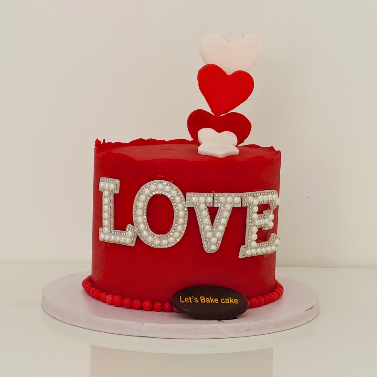 A luxury red celebration cake with 'LOVE' written in pearl-detailed lettering, featuring red and white heart toppers by Let's Bake Cake Abu Dhabi.