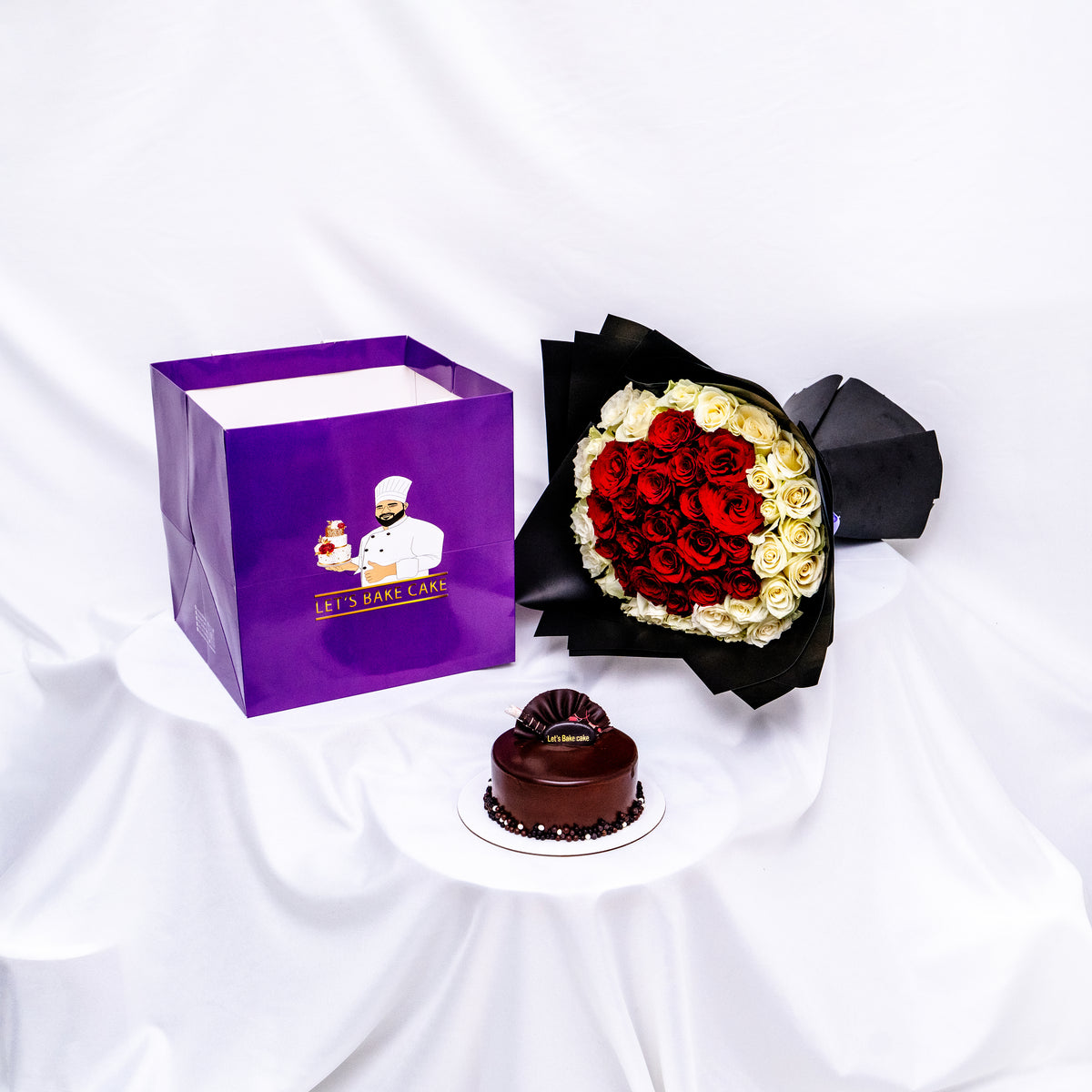 A luxury gift set featuring a rich chocolate glazed bento cake, a heart-shaped red and white rose bouquet in black premium wrap, and a branded 'Let's Bake Cake' purple gift bag, Abu Dhabi.