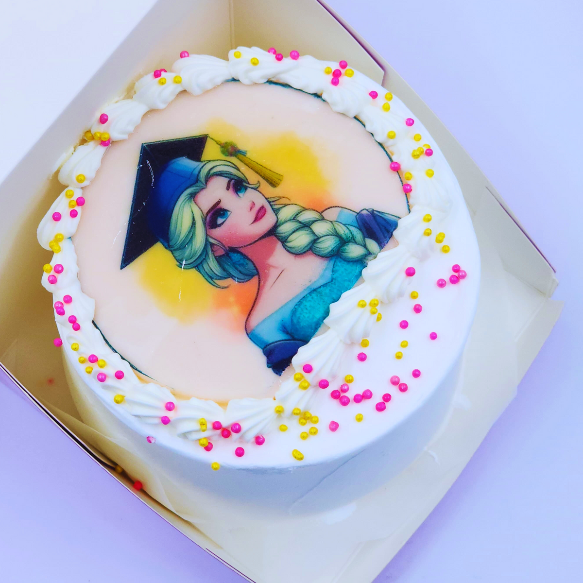 A white graduation cake with an Elsa Frozen edible image, white frosting swirls, and pink/gold sprinkles by Let's Bake Cake Abu Dhabi.