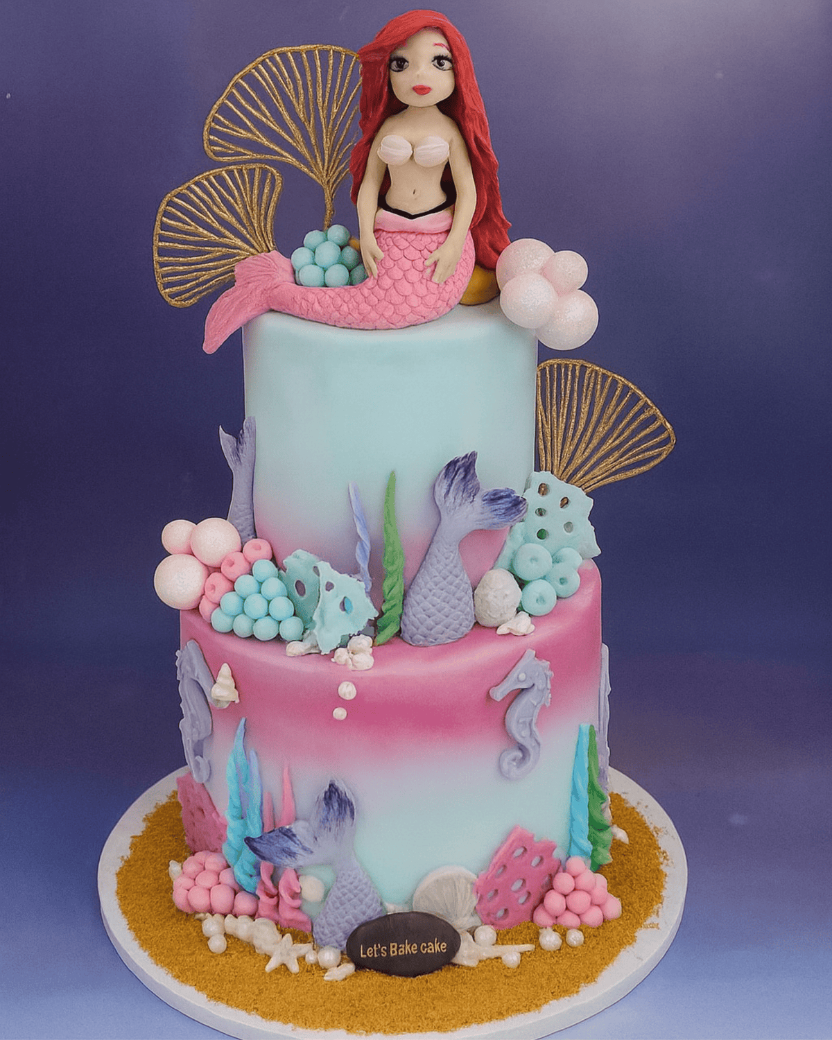 A luxury two-tiered Mermaid themed birthday cake with Ariel figurine, gold sea shells, and colorful coral decorations by Let's Bake Cake Abu Dhabi.