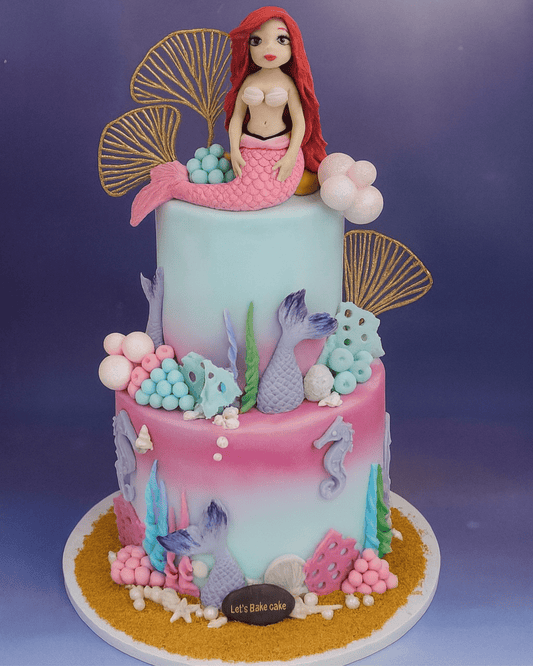 A luxury two-tiered Mermaid themed birthday cake with Ariel figurine, gold sea shells, and colorful coral decorations by Let's Bake Cake Abu Dhabi.