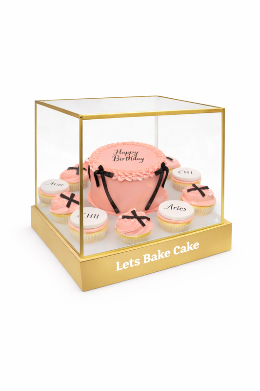A luxury pink birthday cake with black bows and personalized cupcakes inside a gold-framed transparent acrylic box by Let's Bake Cake Abu Dhabi