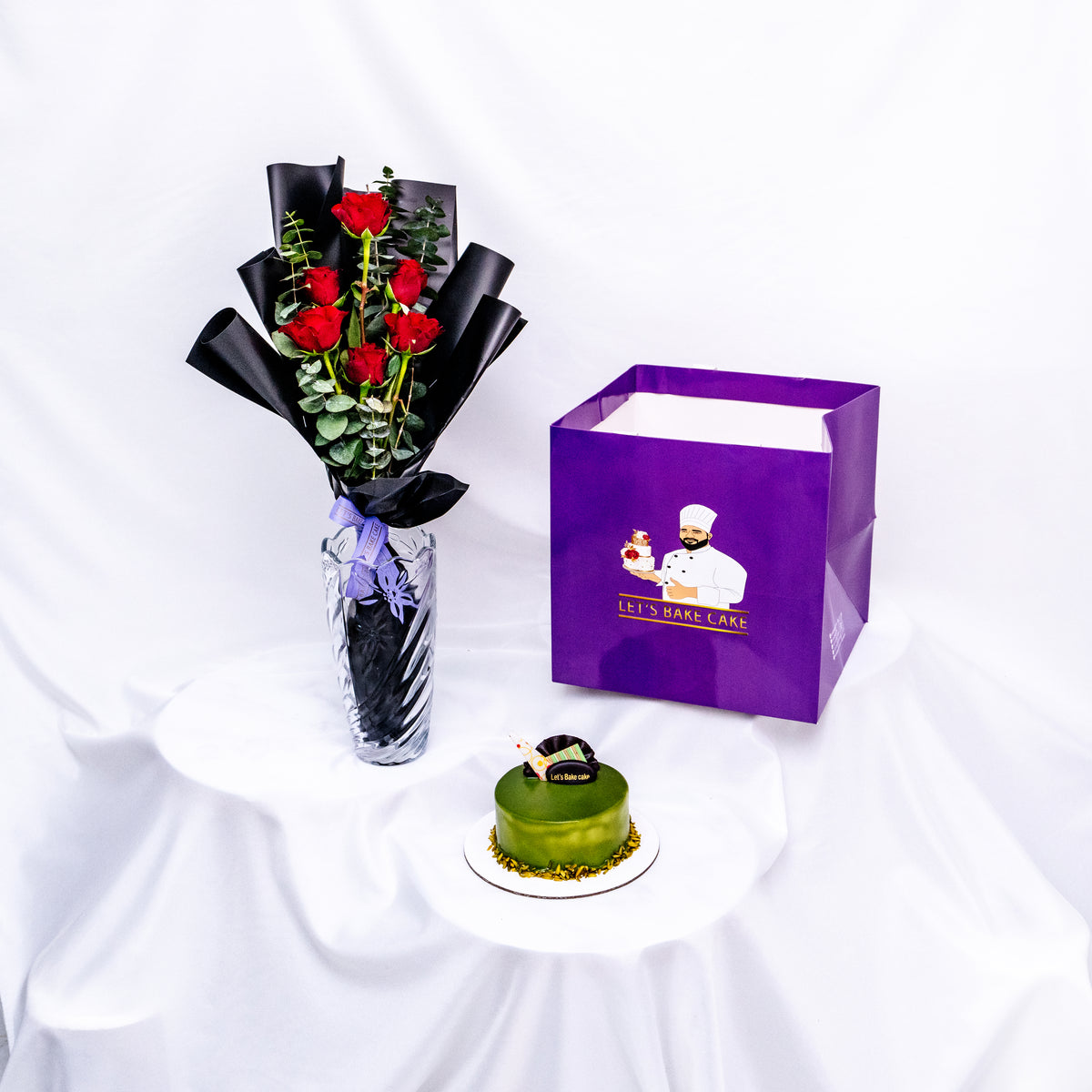 A luxury gift combo featuring a green pistachio glazed bento cake, a premium glass vase with long-stemmed red roses, and a signature 'Let's Bake Cake' purple gift bag, Abu Dhabi.