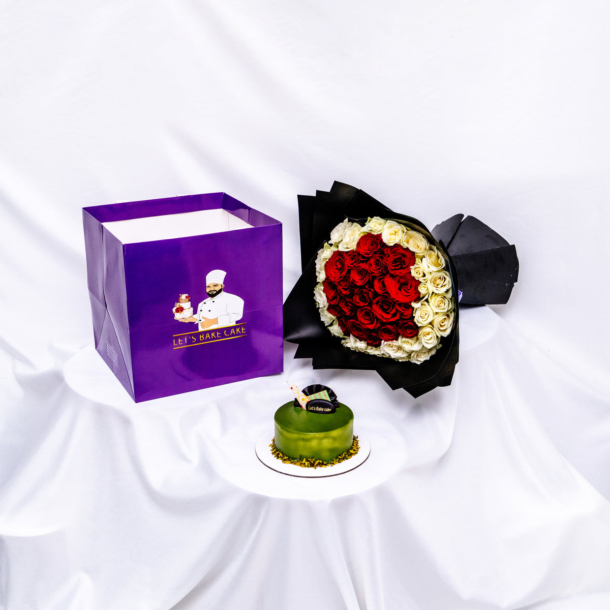 A luxury gift set featuring a green pistachio glazed bento cake, a heart-shaped red and white rose bouquet in premium black wrap, and a branded 'Let's Bake Cake' purple gift bag, Abu Dhabi.