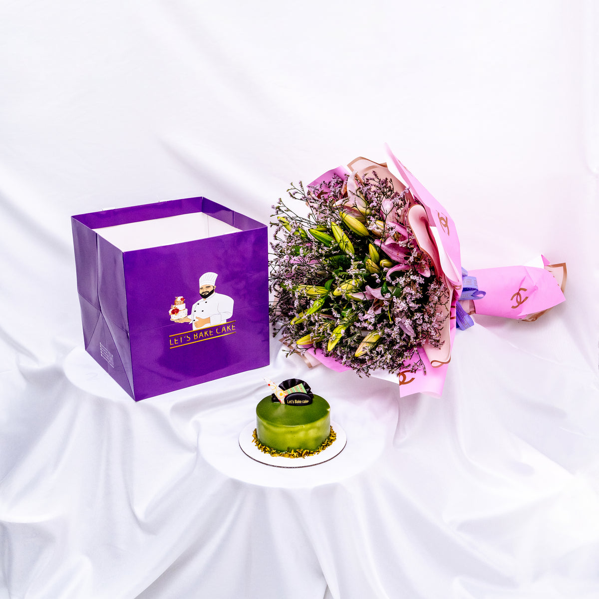 A luxury gift set featuring a green pistachio glazed bento cake, a large pink lily bouquet in designer wrap, and a signature 'Let's Bake Cake' purple gift bag on white silk, Abu Dhabi.