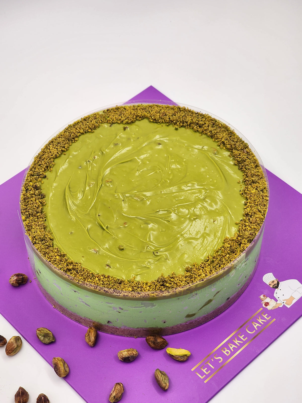 A luxury pistachio cream cake with a smooth green glaze and crushed pistachio border on a purple Let's Bake Cake board.