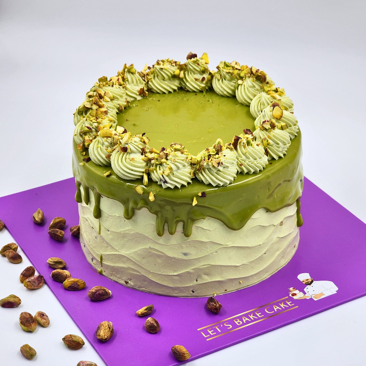 A luxury pistachio drip cake with textured green frosting and crushed pistachios on a purple Let's Bake Cake board