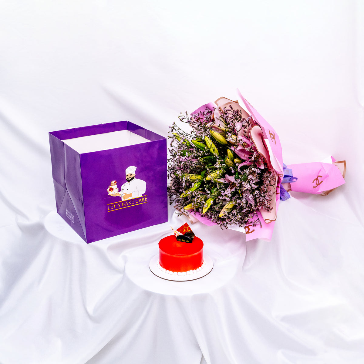 A luxury gift set featuring a vibrant red mirror-glazed bento cake, a large pink lily bouquet in designer wrap, and a signature 'Let's Bake Cake' purple gift bag on white silk, Abu Dhabi.