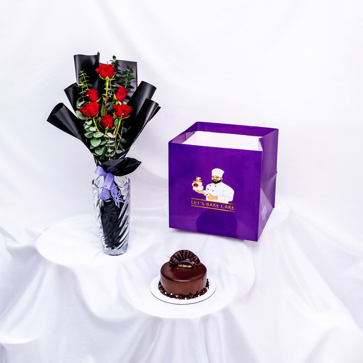 A luxury gift combo featuring long-stemmed red roses in a glass vase, a rich chocolate glazed bento cake, and a signature 'Let's Bake Cake' purple gift bag, Abu Dhabi.