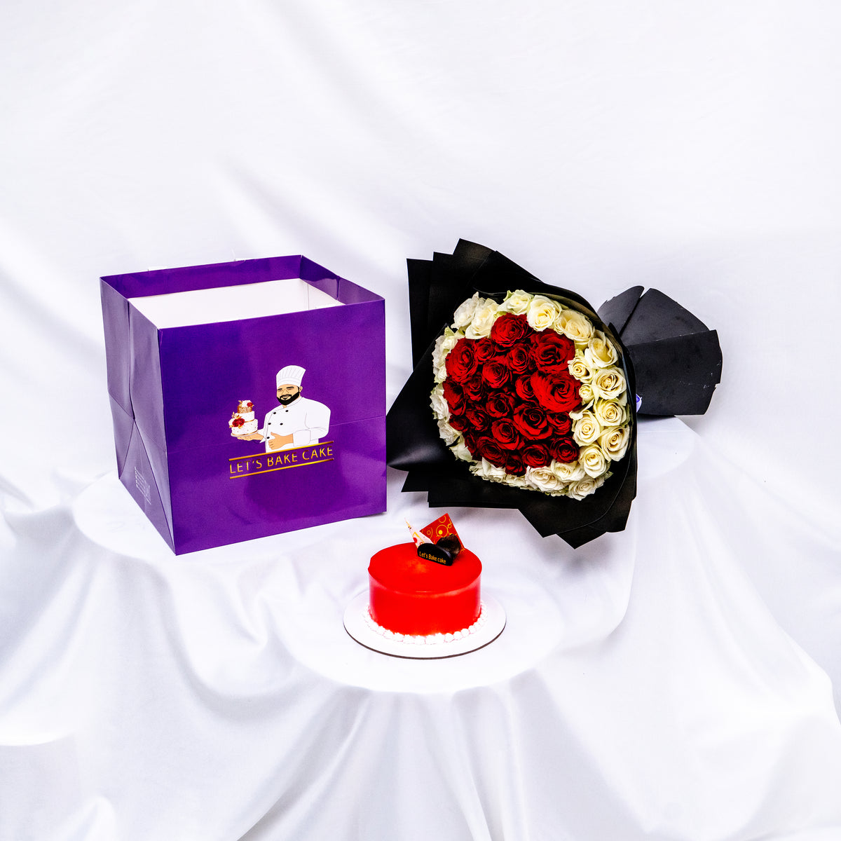 A luxury romantic gift set featuring a vibrant red glazed bento cake, a heart-shaped red and white rose bouquet in black premium wrap, and a branded 'Let's Bake Cake' purple gift bag on white silk, Abu Dhabi