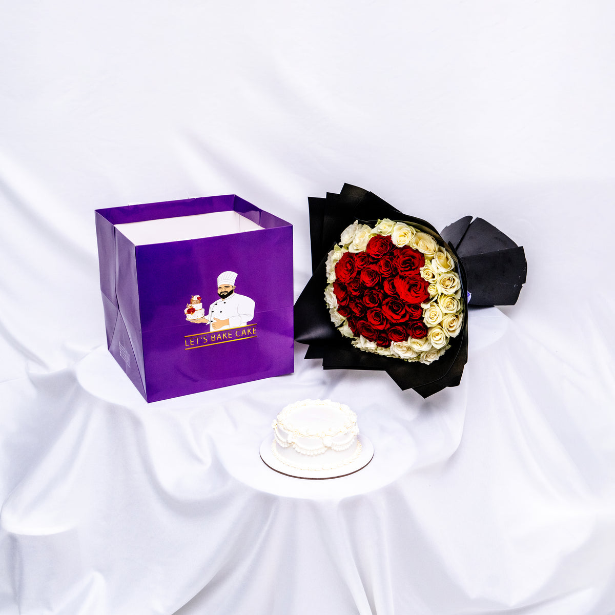 A luxury romantic gift set featuring an elegant white hand-piped bento cake, a heart-shaped red and white rose bouquet in premium black wrap, and a signature 'Let's Bake Cake' purple gift bag, Abu Dhabi.
