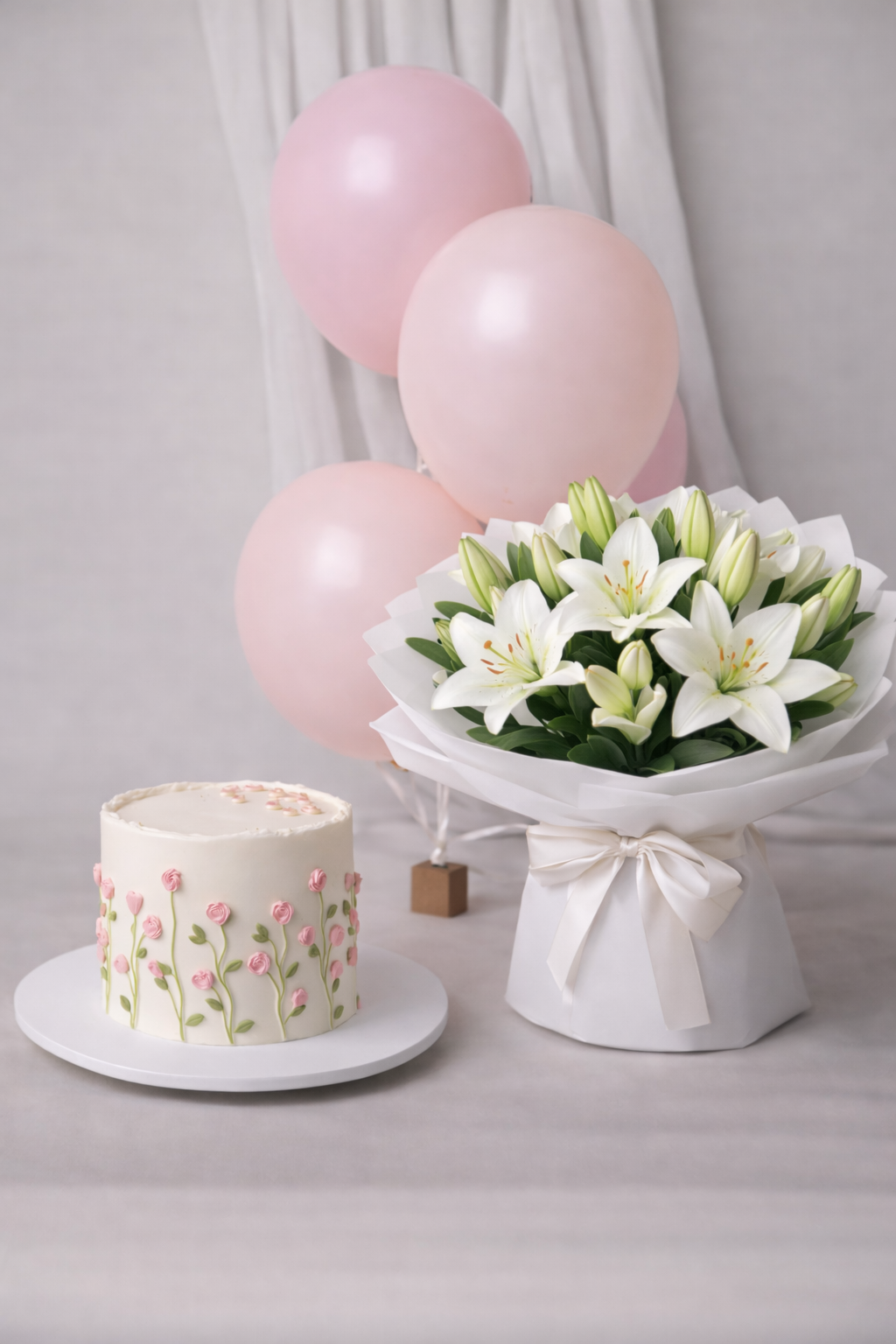 A luxury gift bundle featuring a white cake with pink floral piping, a large bouquet of fresh white lilies, and pink balloons by Let's Bake Cake Abu Dhabi.