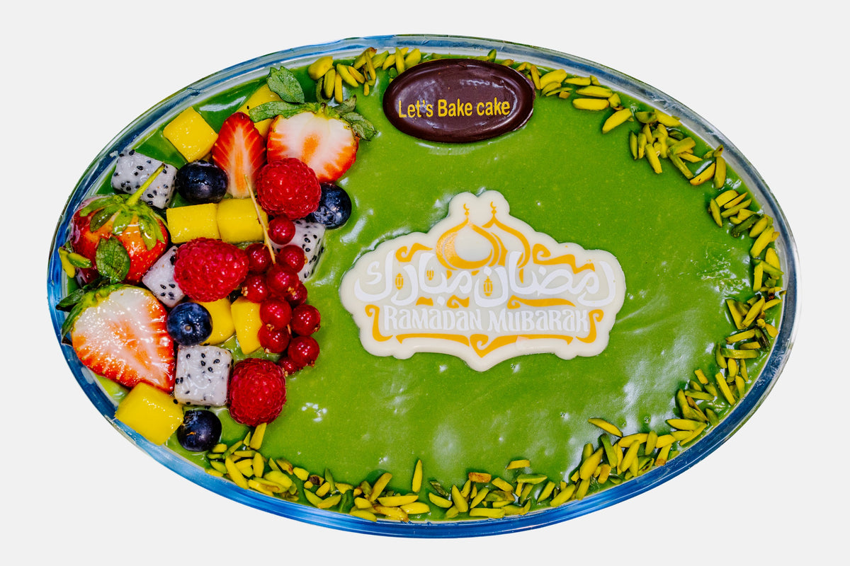 A premium pistachio-glazed fruit cake in an oval glass dish, topped with fresh strawberries, mango, blueberries, and dragon fruit, featuring a 'Ramadan Mubarak' chocolate topper by Let's Bake Cake Abu Dhabi.
