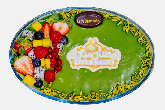 A premium pistachio-glazed fruit cake in an oval glass dish, topped with fresh strawberries, mango, blueberries, and dragon fruit, featuring a 'Ramadan Mubarak' chocolate topper by Let's Bake Cake Abu Dhabi.