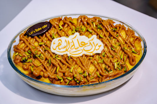 A traditional Ramadan pudding with Biscoff drizzle and pistachios in a glass dish, featuring a festive Arabic chocolate topper by Let's Bake Cake, Abu Dhabi.