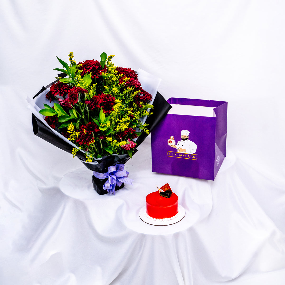 A premium gift set featuring a vibrant red mirror-glazed bento cake, a bold red and yellow flower bouquet in black wrap, and a signature 'Let's Bake Cake' purple gift bag on white silk, Abu Dhabi