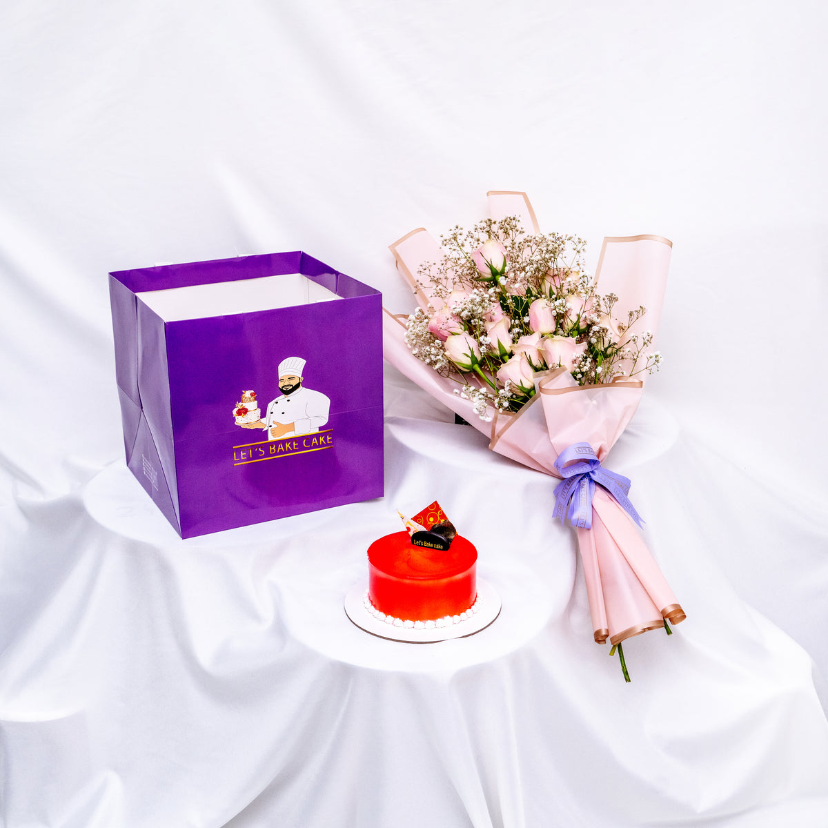 A luxury gift set featuring a vibrant red mirror-glazed bento cake, an elegant pink rose bouquet, and a signature 'Let's Bake Cake' purple gift bag on a white silk backdrop, Abu Dhabi.