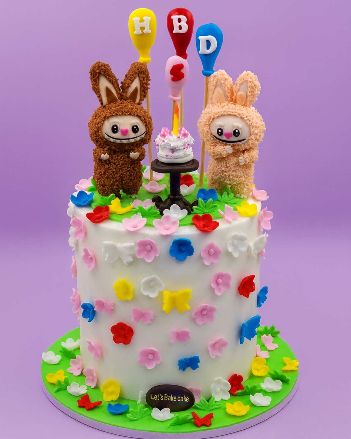A white birthday cake decorated with colorful fondant flowers, butterflies, and two 3D textured bunny characters by Let's Bake Cake Abu Dhabi.